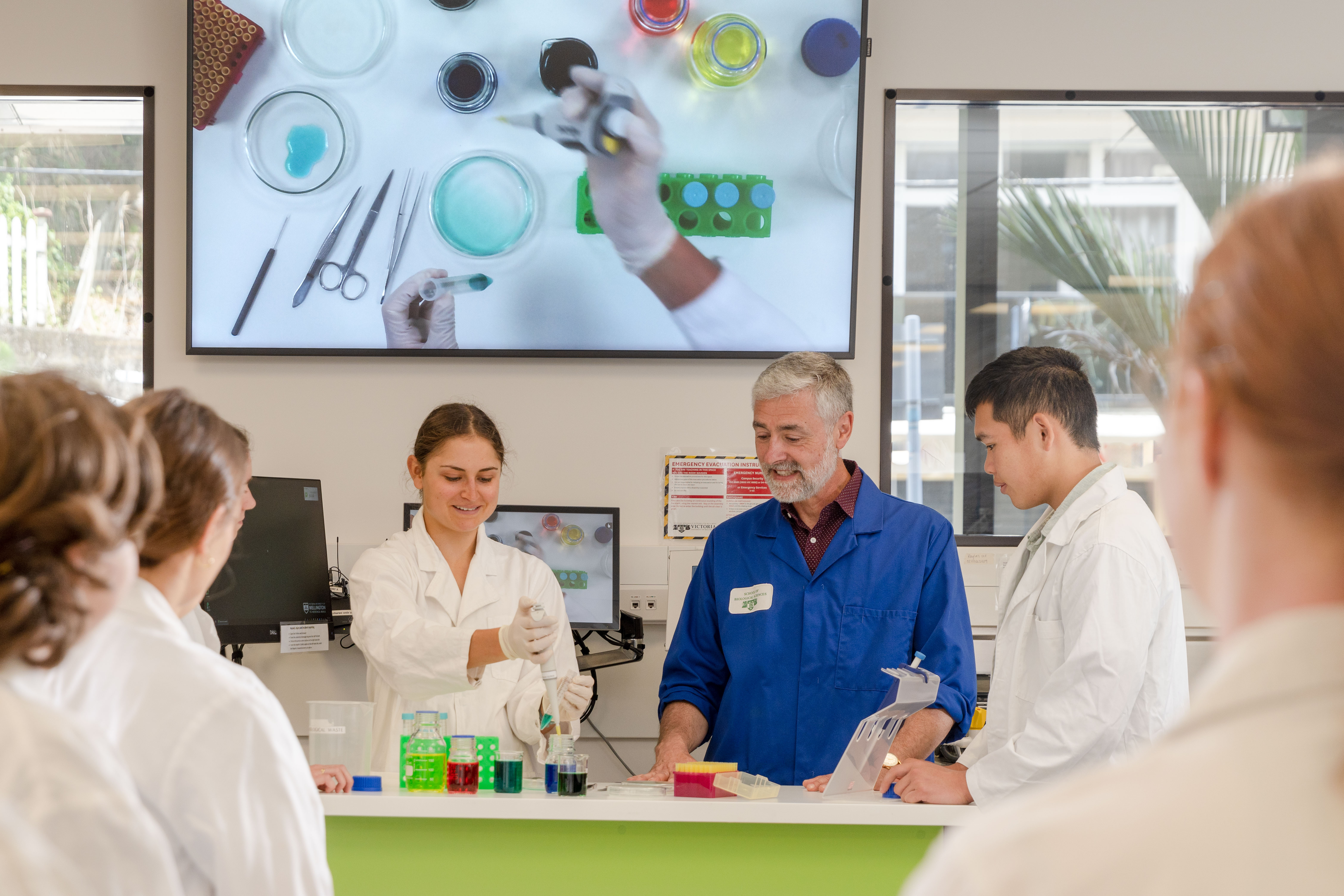 Students and a lecturer in a Biomedical Sciences teaching laboratory observe a pipetting demonstration at a laboratory bench. Coloured solutions, test tubes, and laboratory equipment are arranged on the bench, while a large screen above displays a close-up view of the experiment.