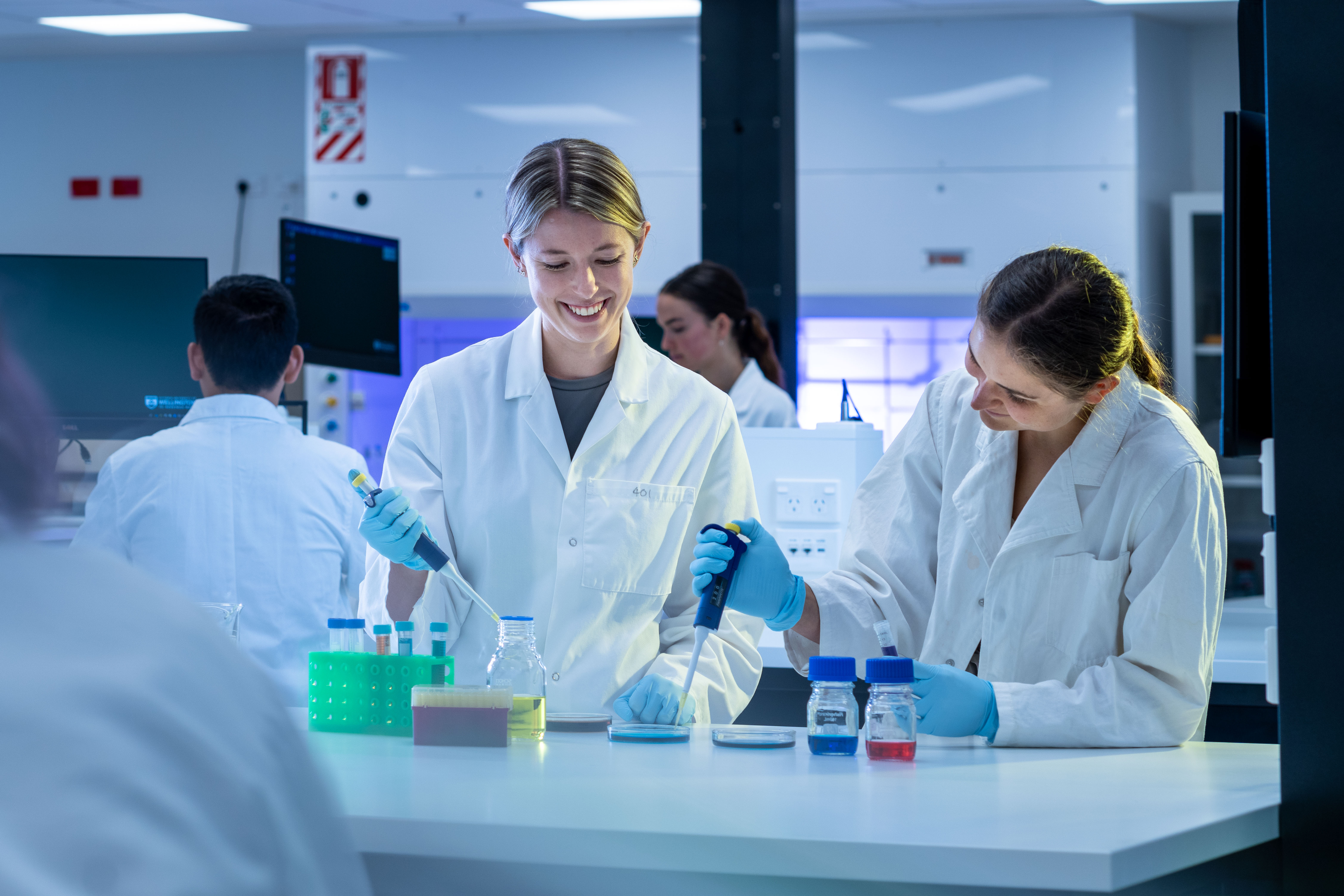 Biomedical Sciences students wearing laboratory coats and gloves use micropipettes to transfer samples in a modern teaching laboratory. Sample tubes, petri dishes, and coloured solutions are arranged on the laboratory bench, while other students work at nearby workstations in the background.