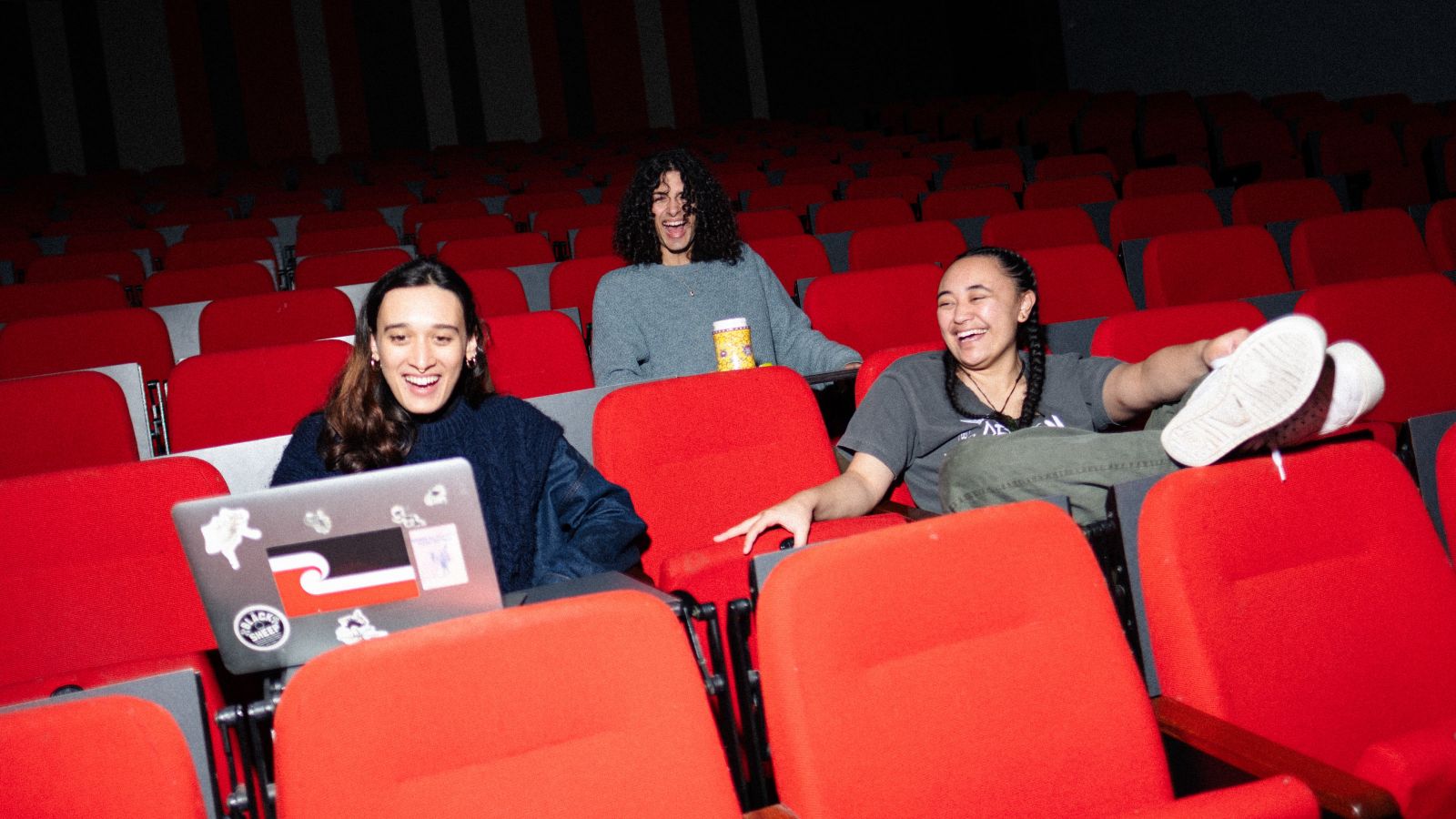 3 students sitting in folding lecture theatre chairs, one has their feet up and all are joking with eachother