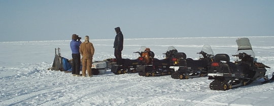 three people and four stationary ski-doos on flat snowy plain