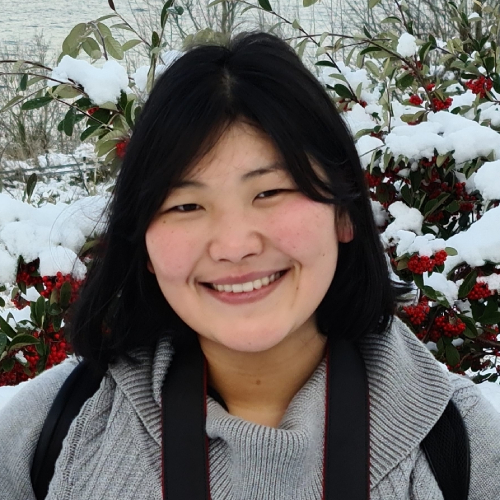 Physics PhD student Angela Xue pictured wearing a light grey knit sweater, smiling in front of a snow covered bush of red berries.