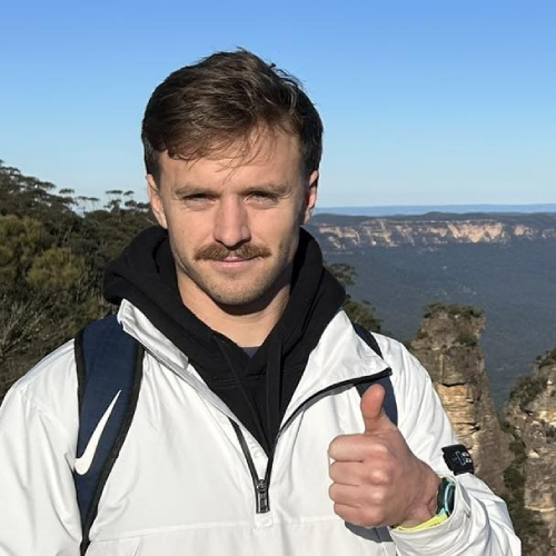 PhD student Finnian Smith holding a 'thumbs up' to camera in front of a blue sky.