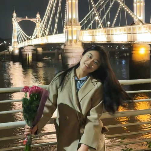Chemistry PhD student Jiaqi Guo, pictured in a tan coat holding a bouqet of roses in front of a lit up bridge at night.