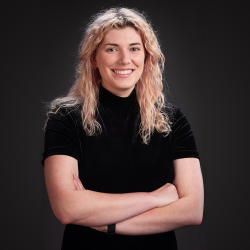 Physics PhD student Mariss Dierkes smiling, wearing a black tshirt with her hair down, crossing her arms in front of a black studio background.