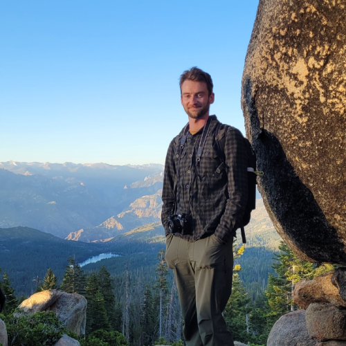 PhD student Mark Bishop standing in front of a boulder, mountains and blue sky in the background.