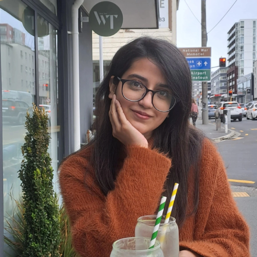 PhD student Qurat Ul Ain is smiling to camera, leaning on her hand, wearing an orange jumper outside a coffee shop.