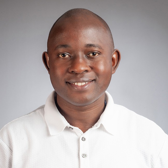 Physics PhD student Viator Mujuni smiling, wearing a white button up shirt in front of a grey studio background.