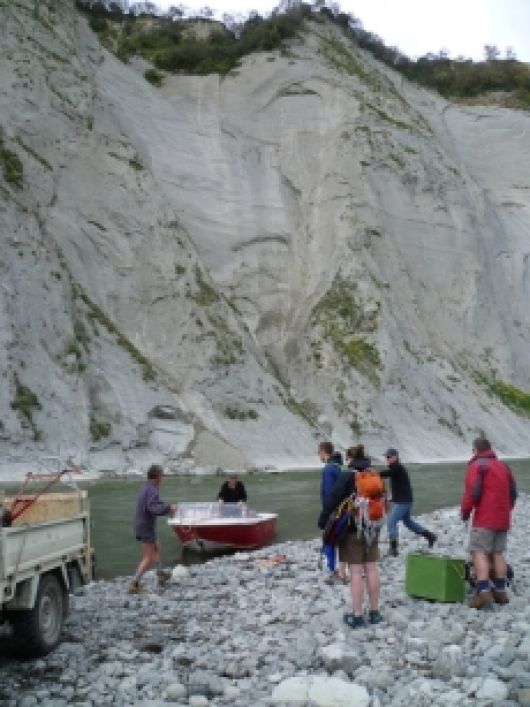 Six people standing on stony riverbank with a motorboat moored on the shore