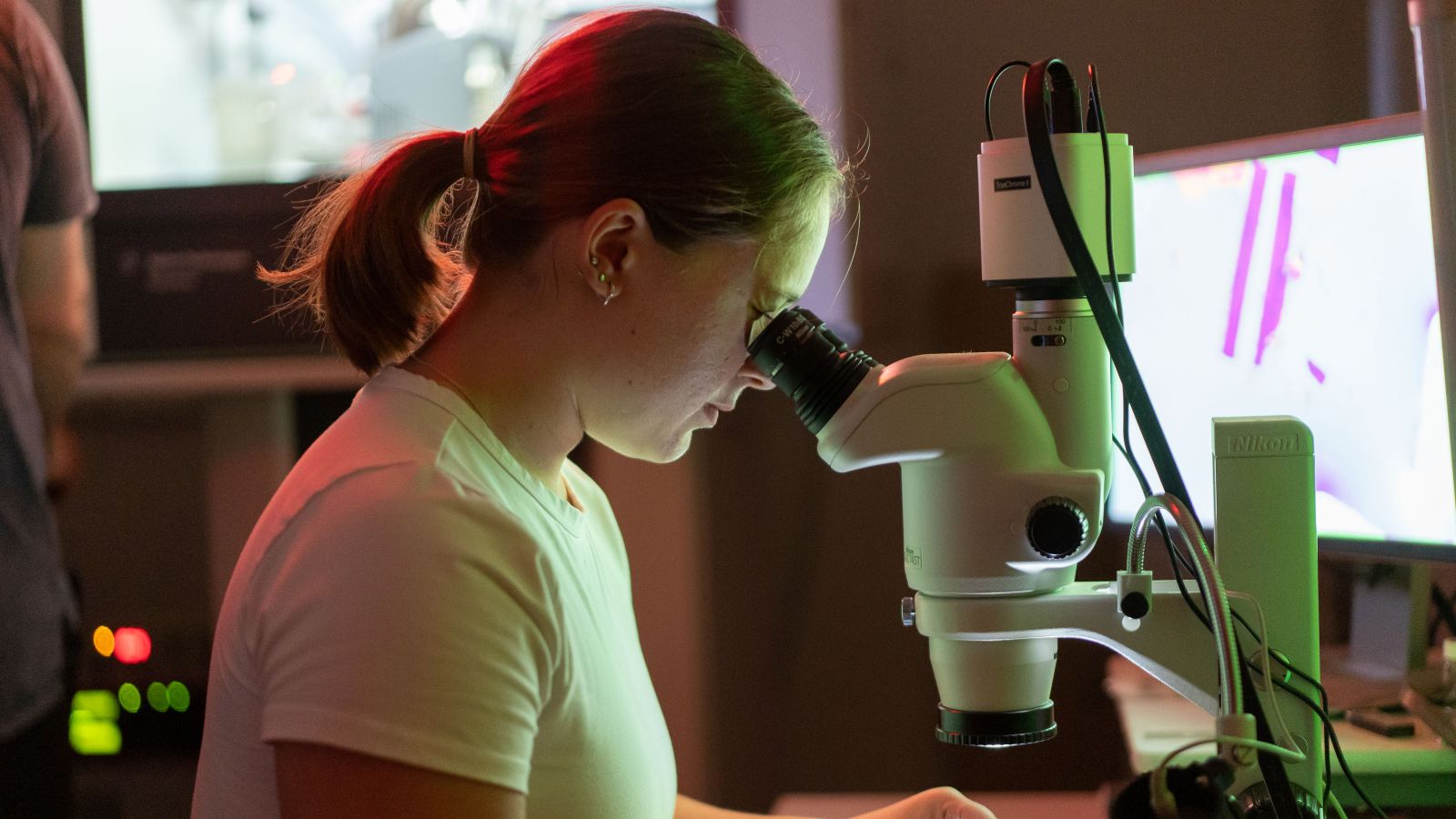 A woman looks into a microscope in a laboratory. Screens are seen in the background.