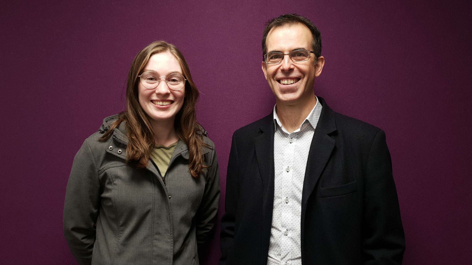 Hayley Mapley and Simon Ferrari stand behind a purple wall smiling to the camera.