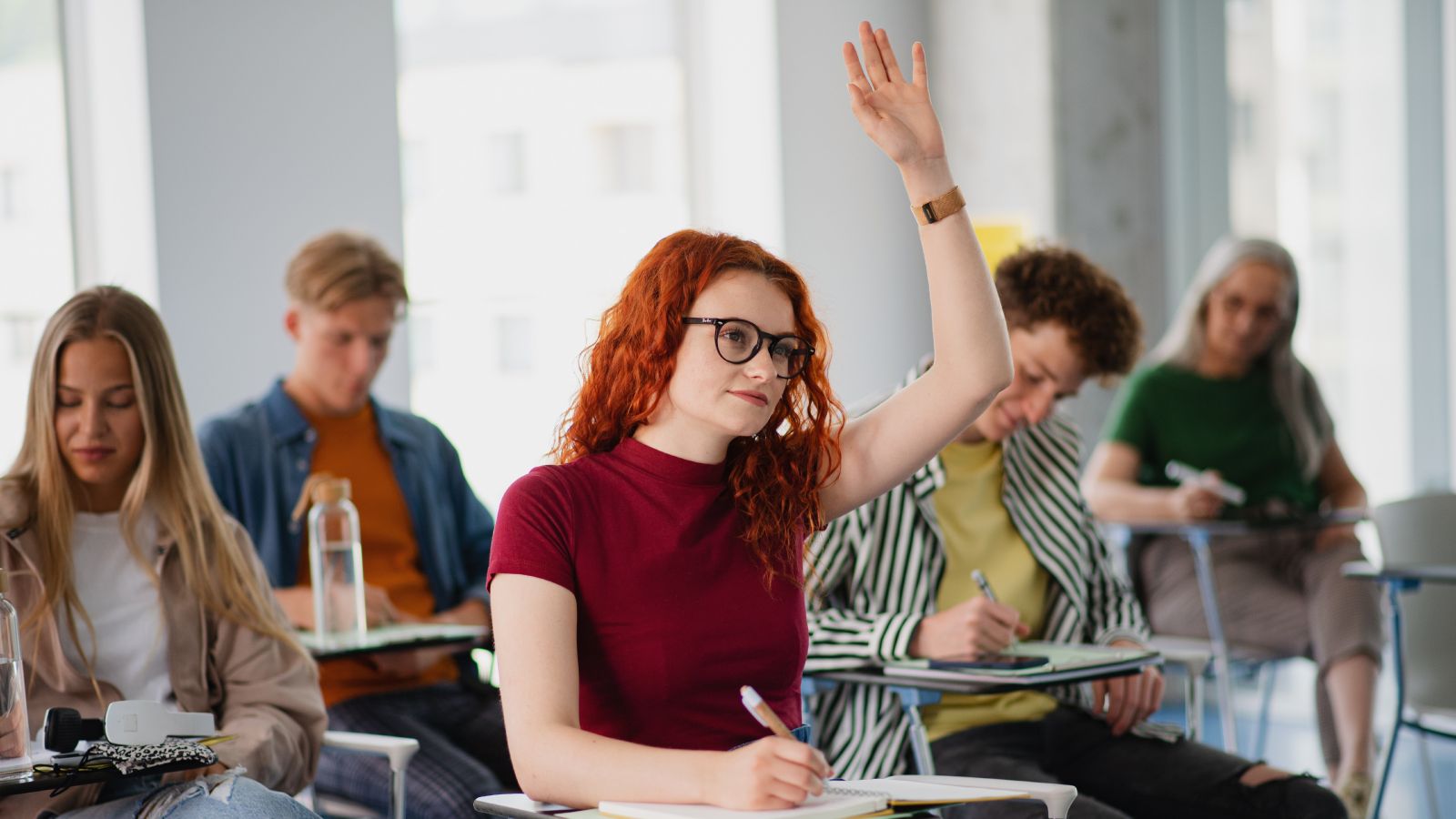 A portrait of a group of university students sitting in a classroom indoors, studying. One has her hand raised to ask a question.