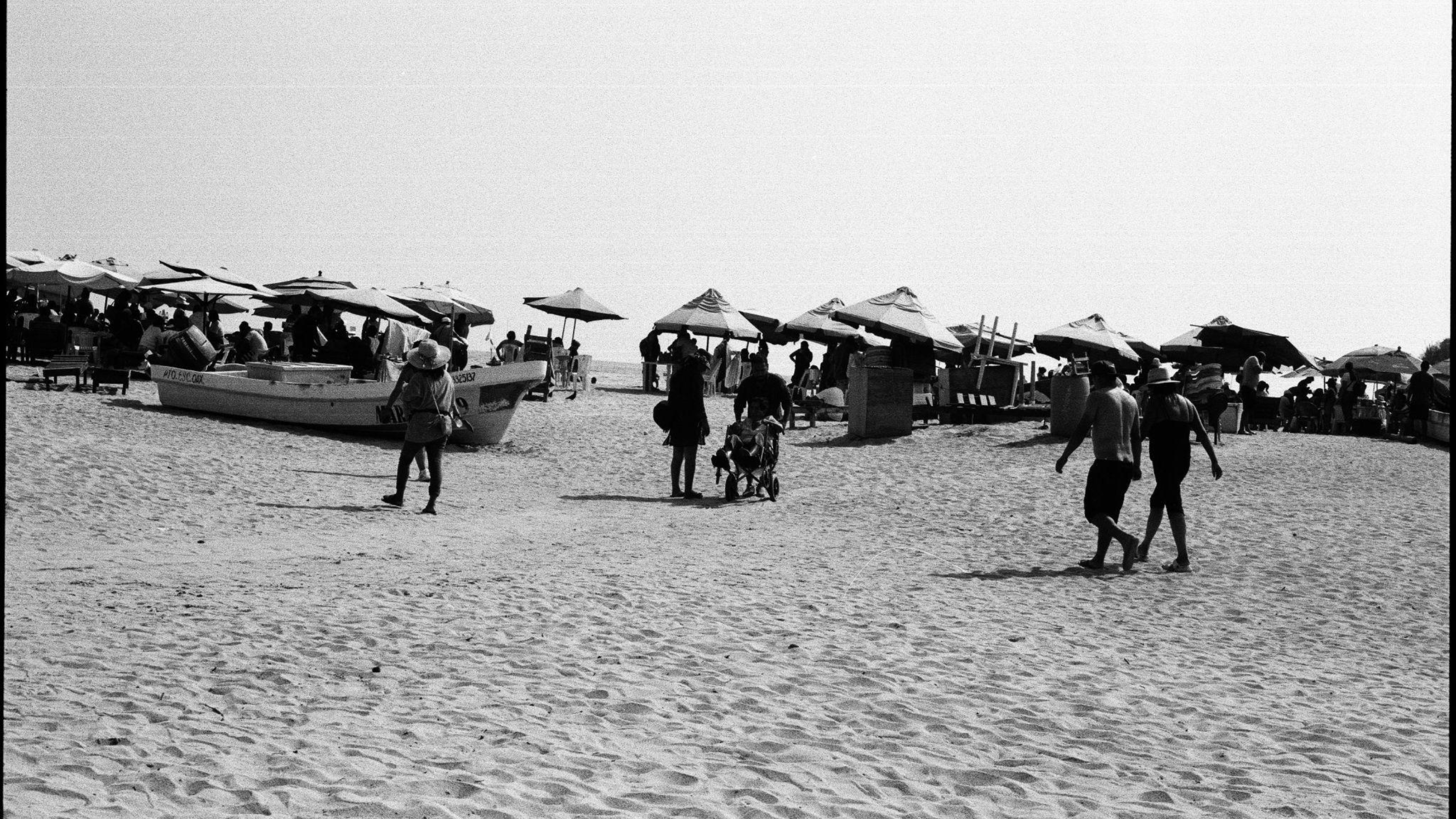 A black and white photo of a beach with many umbrellas visible and people walking around on the sand