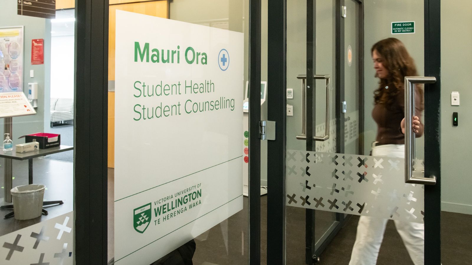 Young female opening door and about to walk inside, with a sign on the door that reads 'Mauri Ora Student Health and Counselling'.