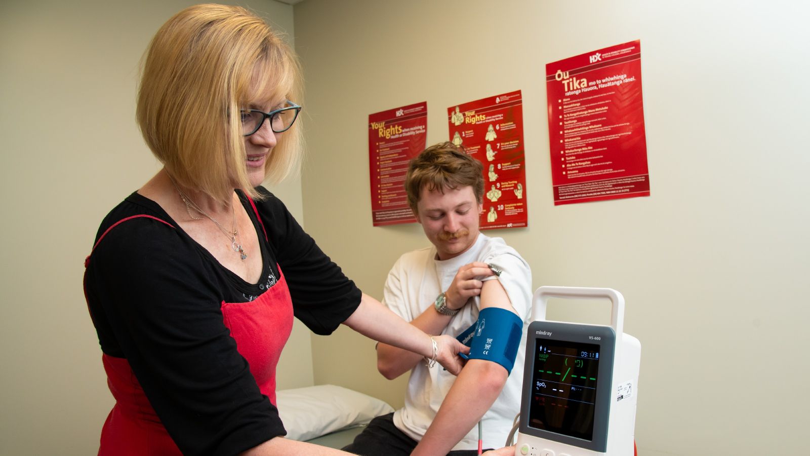 A woman looking at a blood pressure reading on a machine with a young man wearing a blood pressure cuff in the background.