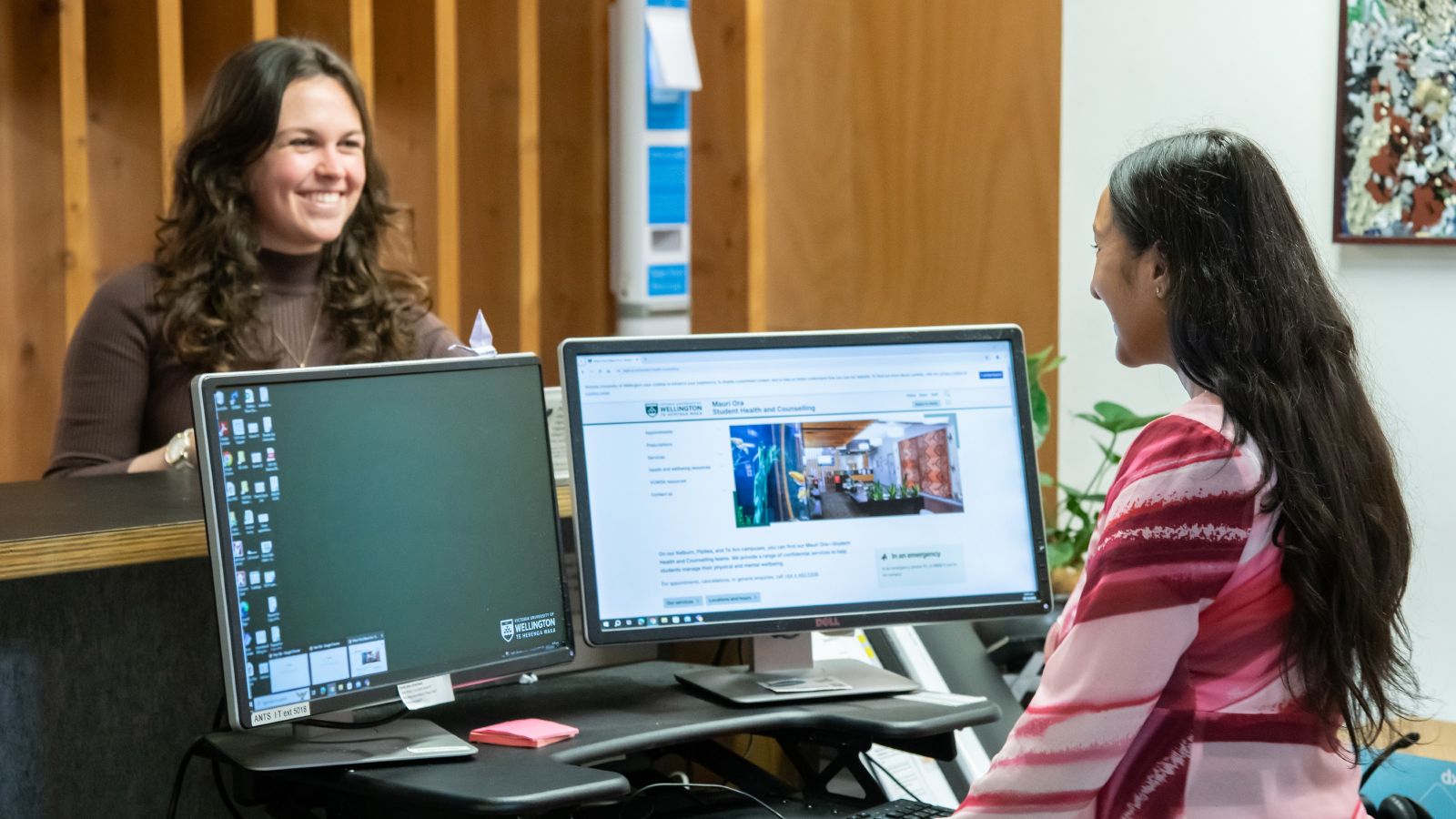 Young female standing behind reception speaking to a female receptionist.