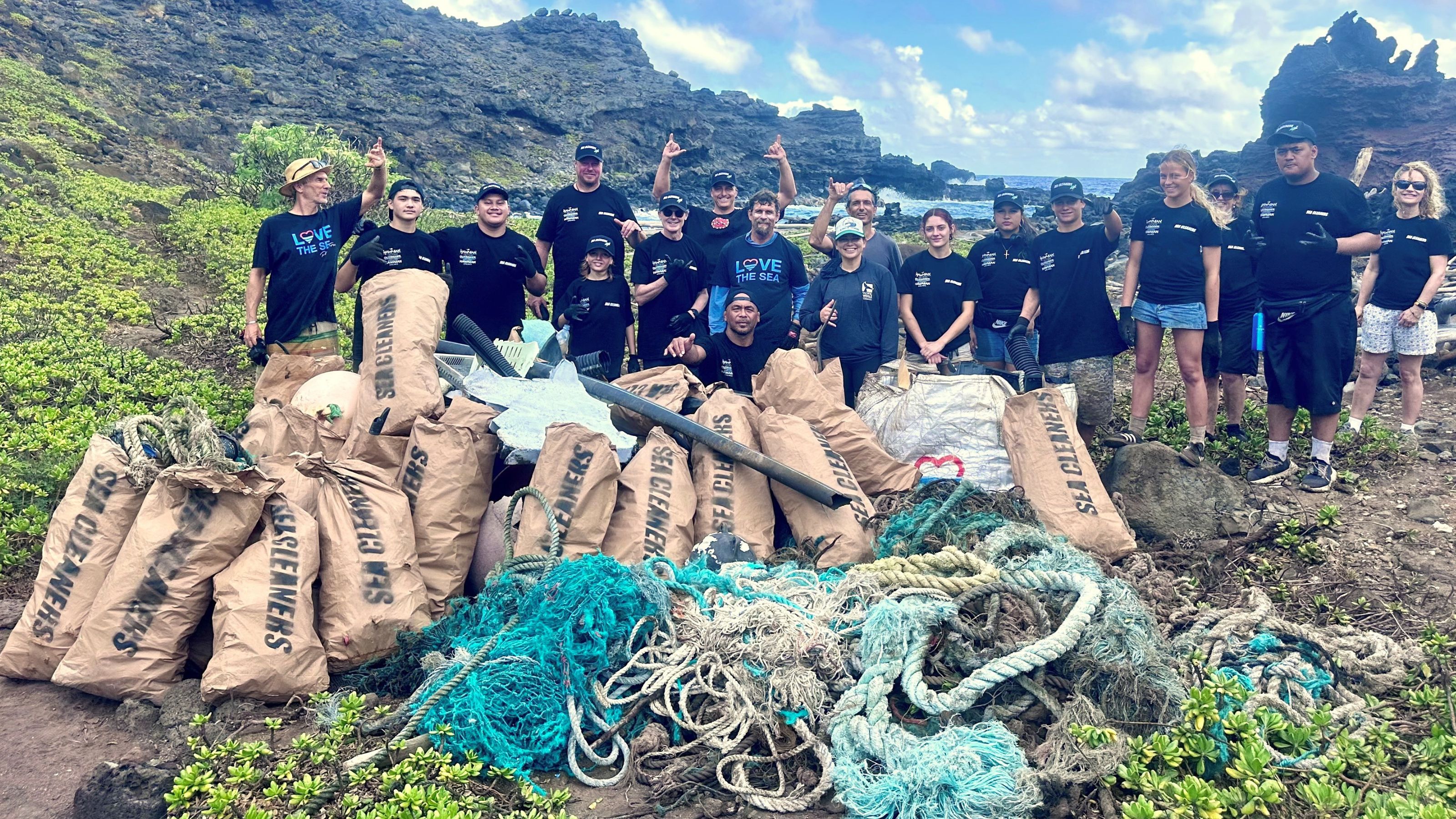A group of about 20 young people stand on a beach triumphantly in front of a dozen full rubbish sacks and nylon netting and rope cleared from the ocean