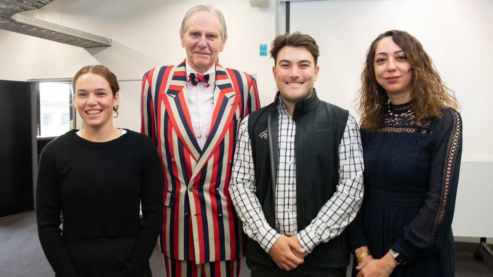 Richard Holden stands next to scholarship recipients Faeze, Hannah and James, wearing a blue, red and white striped suit