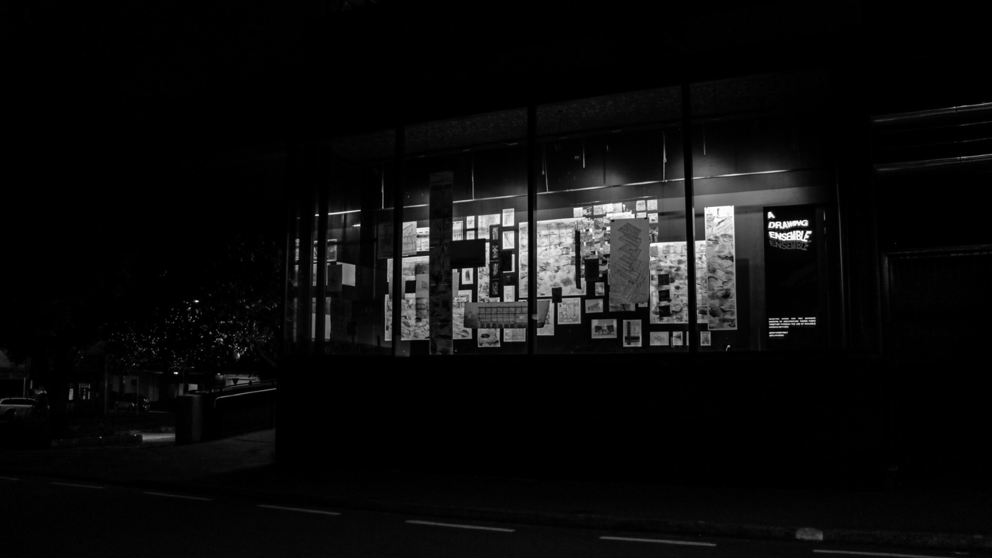 Night shot of the window gallery, black back drop and light shining through form the gallery space showing drawings hang and displayed inside the space 