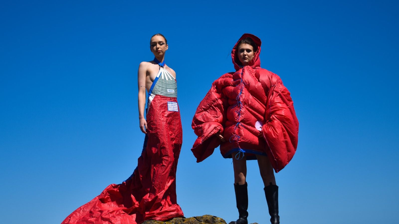 Two models stand on rocks with a blue sky in the background, one wears a red hand made puffer jacket and the other wears a long red gown. Both are made from sailcloth