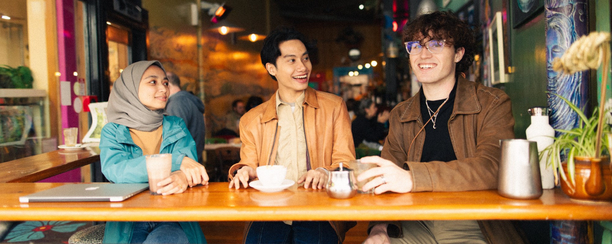 Three students sit by a table in a cafe, drinking coffee and engaged in conversation while looking out.